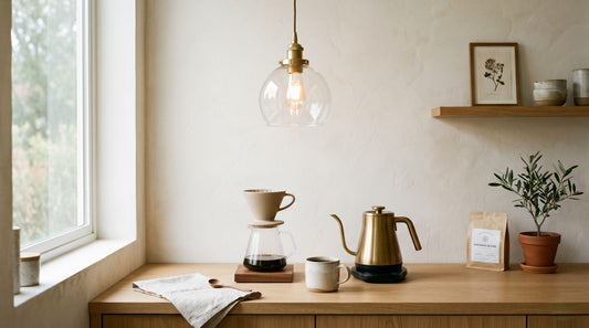 A minimalist home café setup featuring a brass gooseneck kettle and pour-over coffee dripper on a wooden counter, illuminated by a warm glass pendant light against a neutral textured wall.